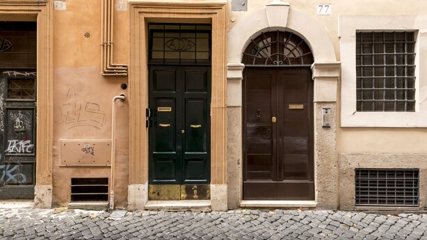 Une façade d’immeuble avec deux portes noires, numérotées 75 et 77, et une fenêtre ornée d’une grille métallique.