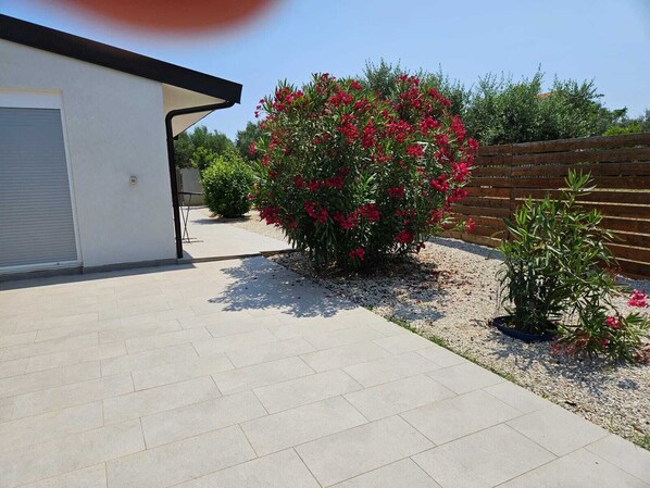 Uma entrada pavimentada leva a uma porta de garagem, cercada por um jardim com plantas floridas vermelhas e uma cerca de madeira.