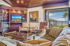 A living room with a large flat-screen TV, a bookshelf, a comfortable sofa, and a view of the ocean through a sliding glass door. - Captivating Tropical Escape at Wailea Elua 2202 - Breathtaking Ocean Views (Wailea, Maui)