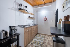 Uma cozinha compacta com fogão a gás, torradeira e cafeteira. Possui armários de madeira, bancada preta e tapete estampado.