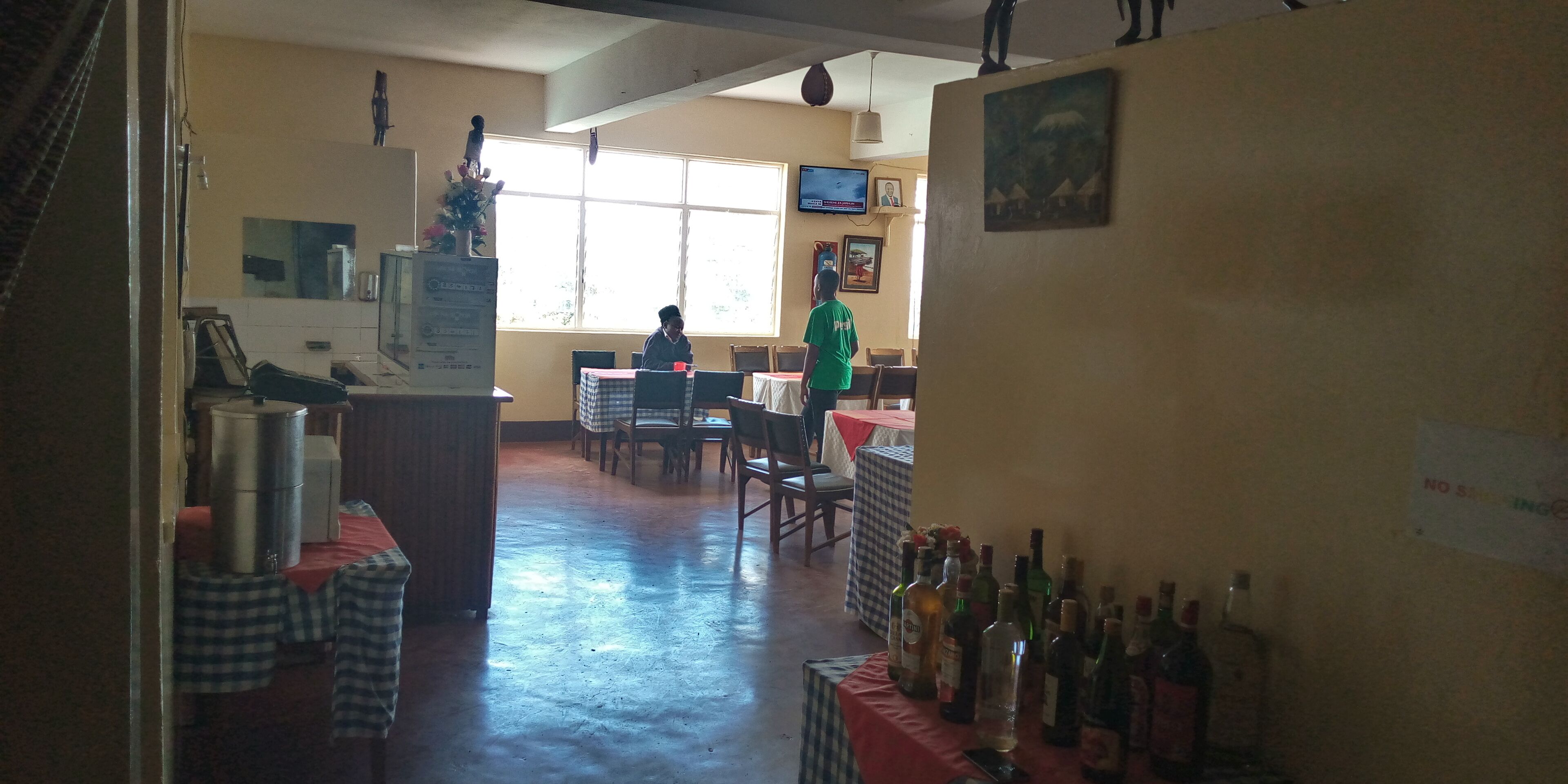 A restaurant with a long counter, tables, and chairs. There are bottles on the counter and a TV on the wall.