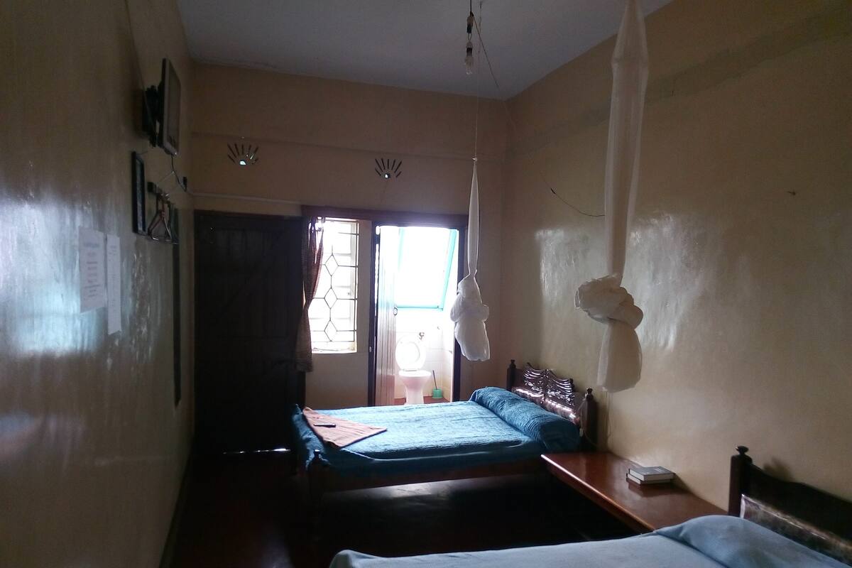 A bedroom with two beds, a wooden headboard, a bedside table, a hanging light fixture, and a window with curtains.