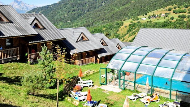A chalet-style hotel with multiple wooden cabins, a glass-enclosed pool area, and a mountainous backdrop. - Rental in the Mountain at Saint François LONGCHAMPS (Saint François LONGCHAMPS)