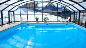 A clear blue pool with a glass ceiling, surrounded by lounge chairs and potted plants, with a snowy mountain view in the background. - Rental in the Mountain at Saint François LONGCHAMPS (Saint François LONGCHAMPS)