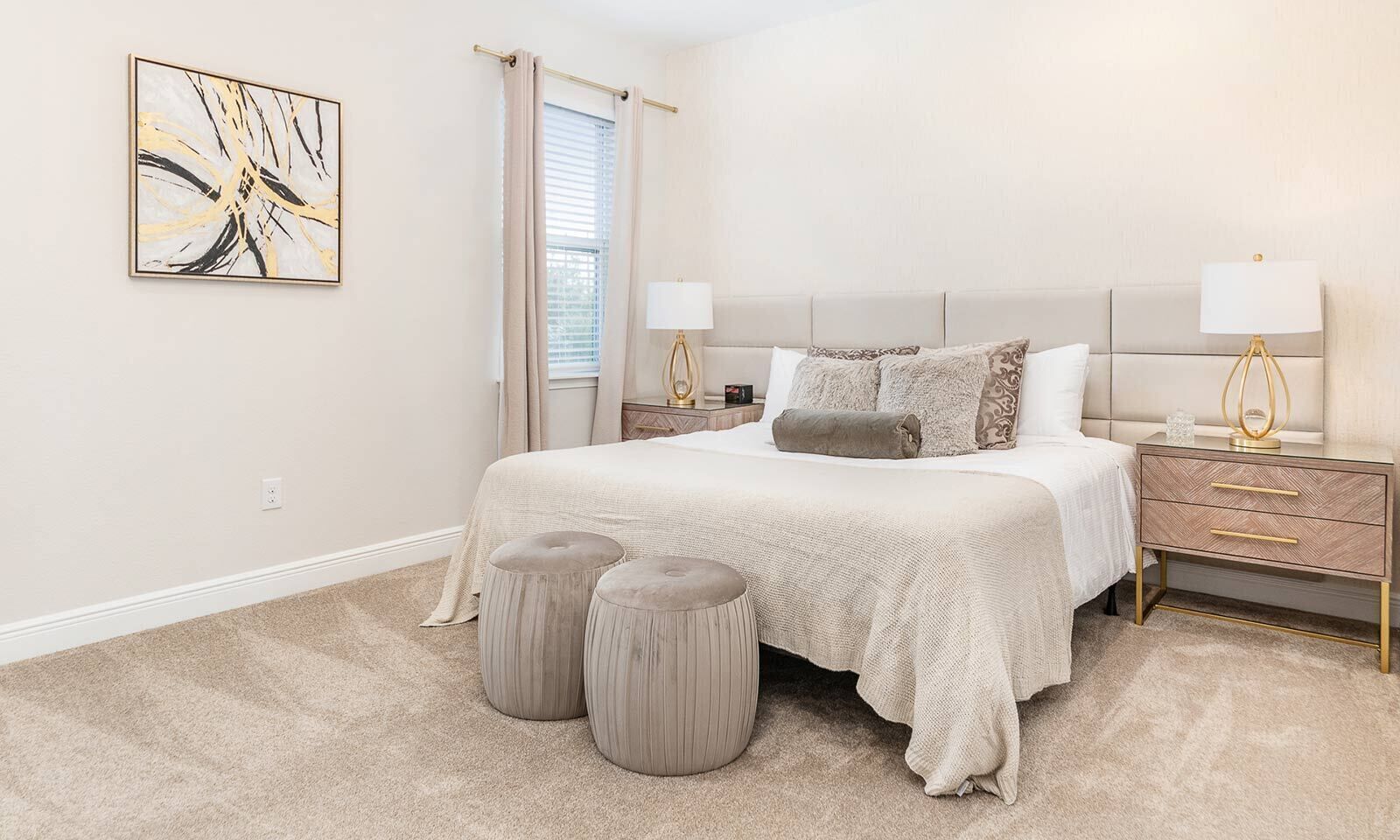A bedroom with a bed, two bedside tables, a nightstand, a window with curtains, and a wall-mounted abstract painting.