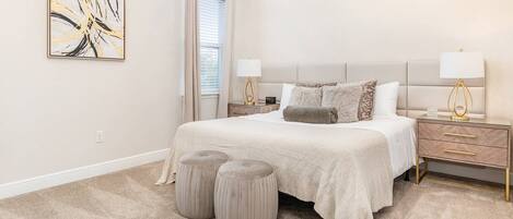 A bedroom with a bed, two bedside tables, a nightstand, a window with curtains, and a wall-mounted abstract painting.