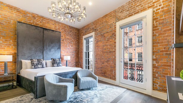 A bedroom with a large bed, two armchairs, a chandelier, and a view of a brick building through a window.