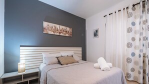 Et moderne soveværelse med en stor seng, sengegavl, et natbord, en lampe og et bybillede på væggen.