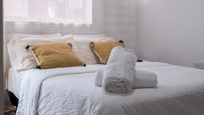 Una cama bien hecha con almohadas blancas y mostaza, una toalla blanca enrollada y una ventana con cortinas traslúcidas.