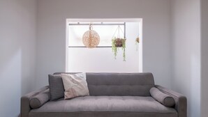 Un sofá gris con un cojín estampado, una lámpara colgante tejida y una planta colgante con hojas colgantes.