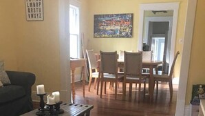 En stue med en mørk sofa, et sofabord med stearinlys, og et træspisebord med stole. Der er et billede på væggen og en loftsvifte over spiseområdet.