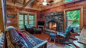 Un interior de cabaña acogedor con chimenea de piedra, paredes de madera y ventanas amplias que dan a un bosque.