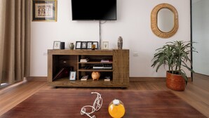 Un salon avec un téléviseur à écran plat fixé au mur, un meuble de rangement en bois contenant divers objets, un grand miroir et une plante en pot.