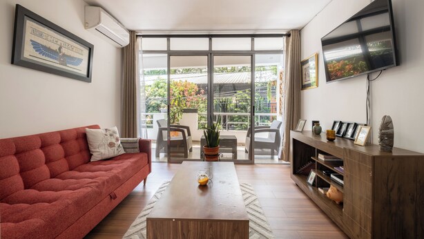 Un salon avec un canapé rouge, une table basse en bois et un téléviseur fixé au mur. Une porte-fenêtre coulissante donne accès à un balcon agrémenté de plantes et de mobilier d’extérieur.