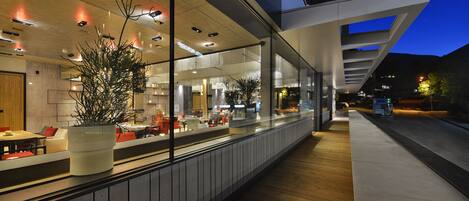 A modern restaurant with a long counter, large potted plants, and a view of the city at night.