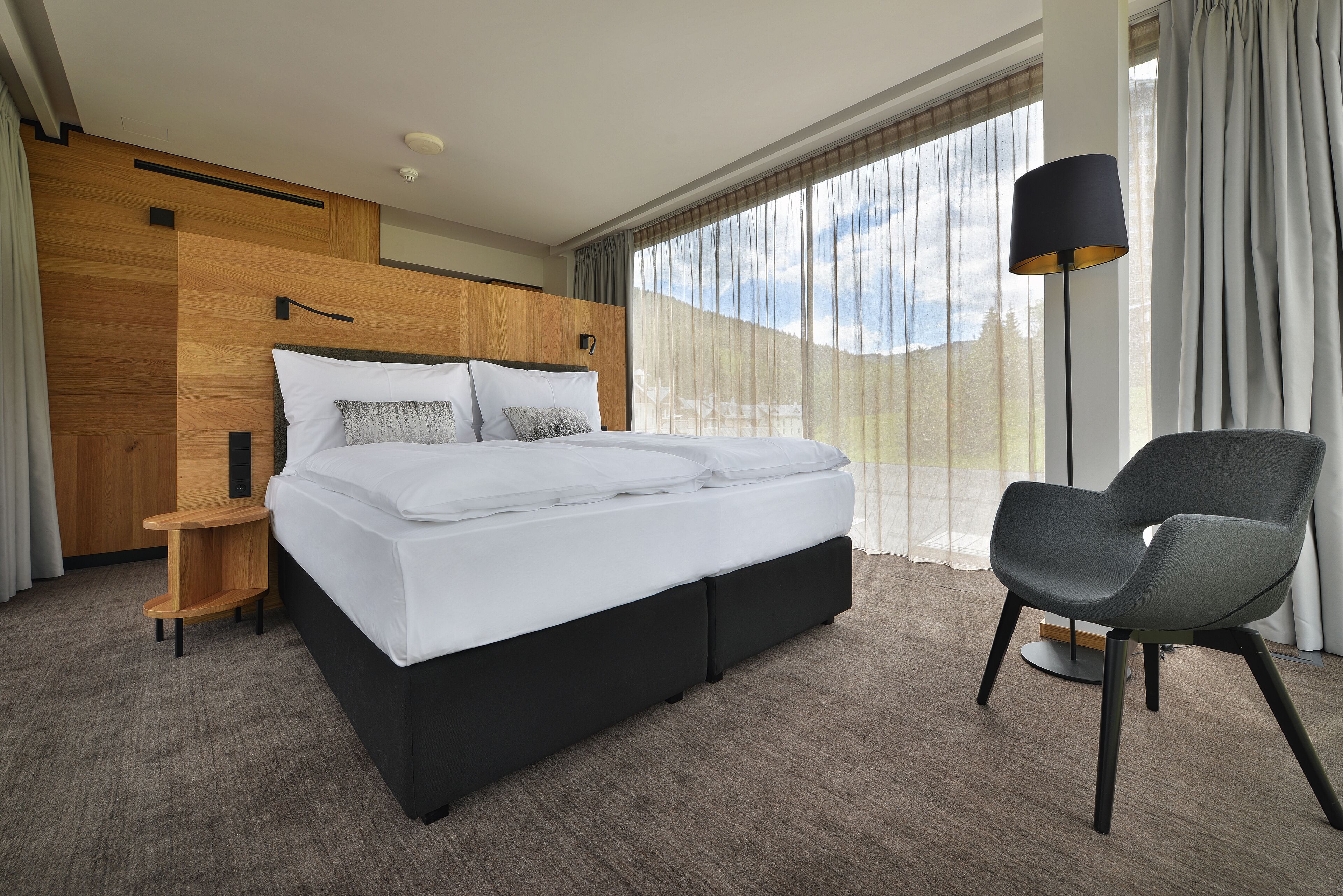 A modern hotel room with a large bed, a wooden headboard, a side table, a chair, and a large window with curtains.