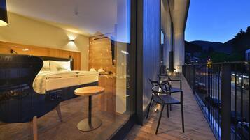A modern hotel room with a large bed, a small table, and a balcony with a view of the city and mountains.