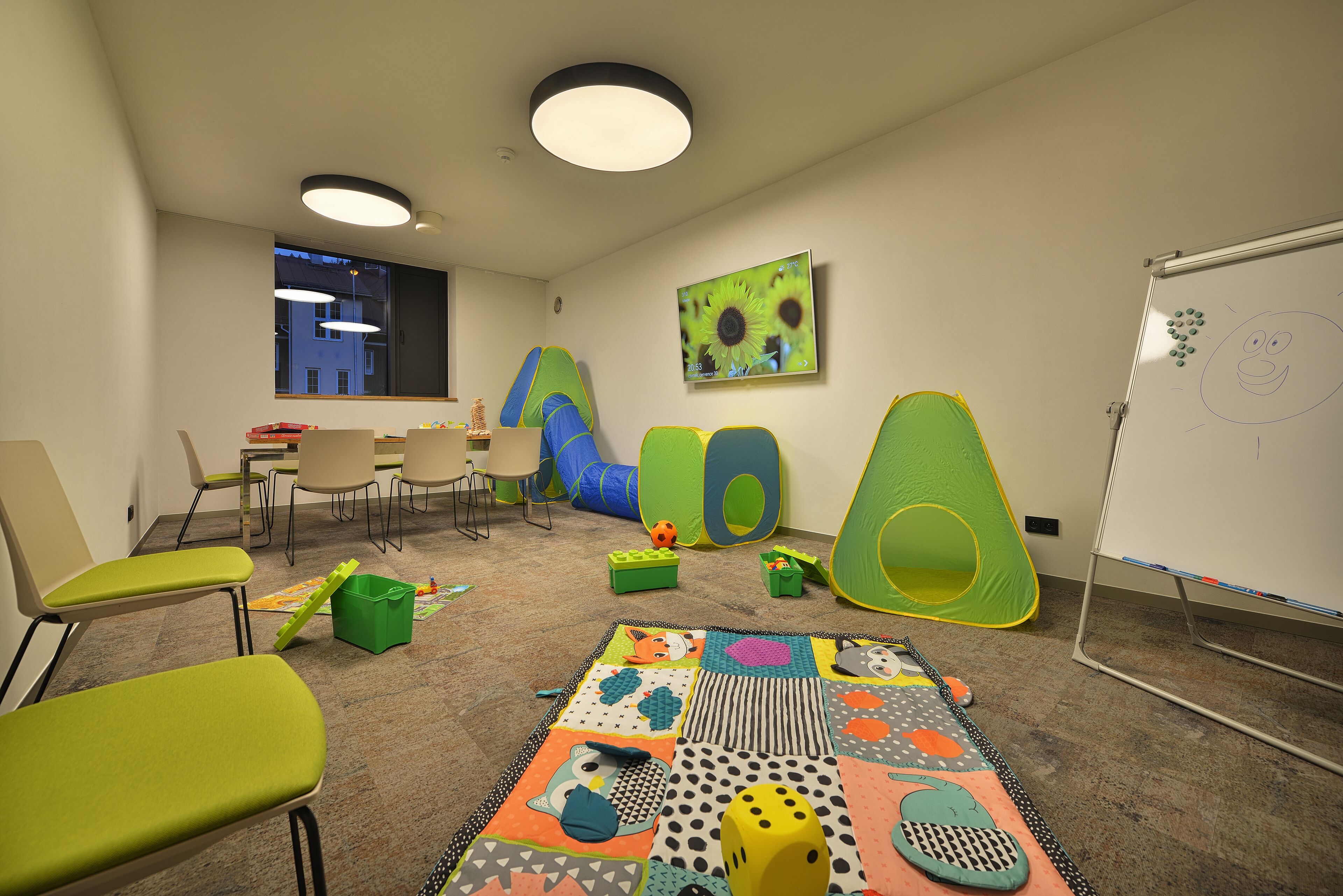A children's playroom with colorful play mats, green and blue play structures, and a whiteboard with drawings.
