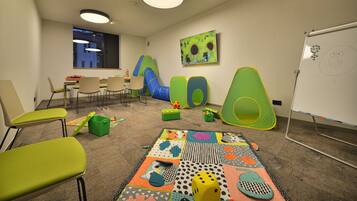 A children's playroom with colorful play mats, green and blue play structures, and a whiteboard with drawings.