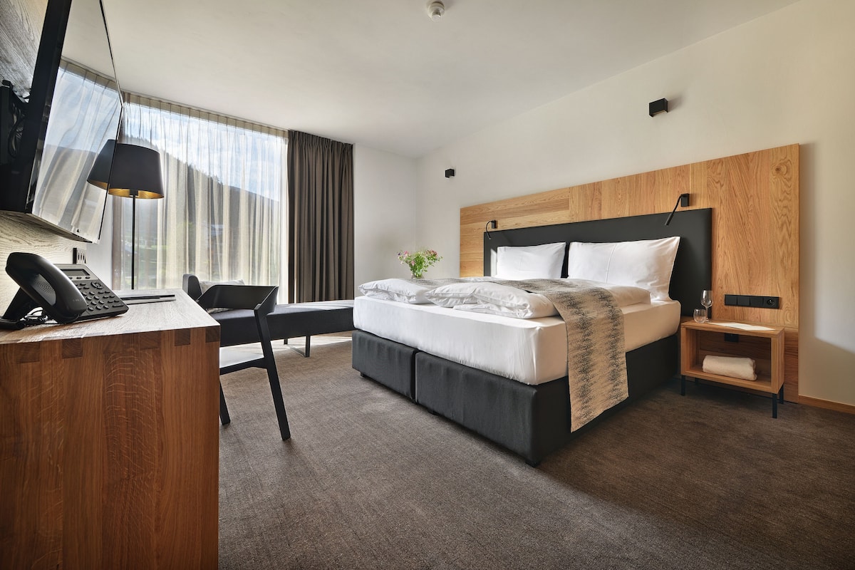 Ein modernes Hotelzimmer mit Schreibtisch aus Holz, einem Bett mit Kopfteil, einem Nachttisch und einem großen Fenster mit Vorhängen.