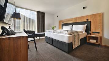 A modern hotel room with a wooden desk, a bed with a headboard, a bedside table, and a large window with curtains.