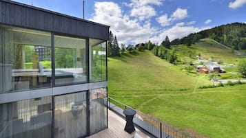 En moderne bygning med store glasvinduer, en træterrasse og udsigt over et grønt landskab med bakker og en lille landsby.