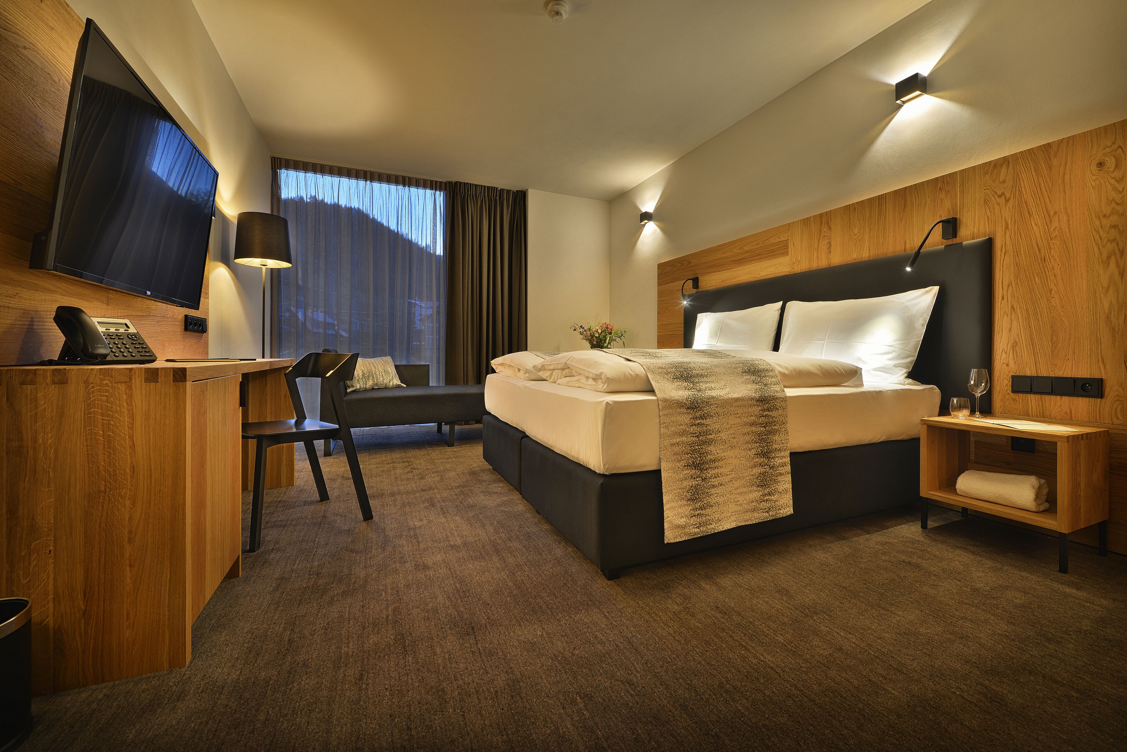 Habitación de hotel moderna con una cama grande, un escritorio con teléfono, un televisor de pantalla plana y una zona de estar con una silla y una mesita.