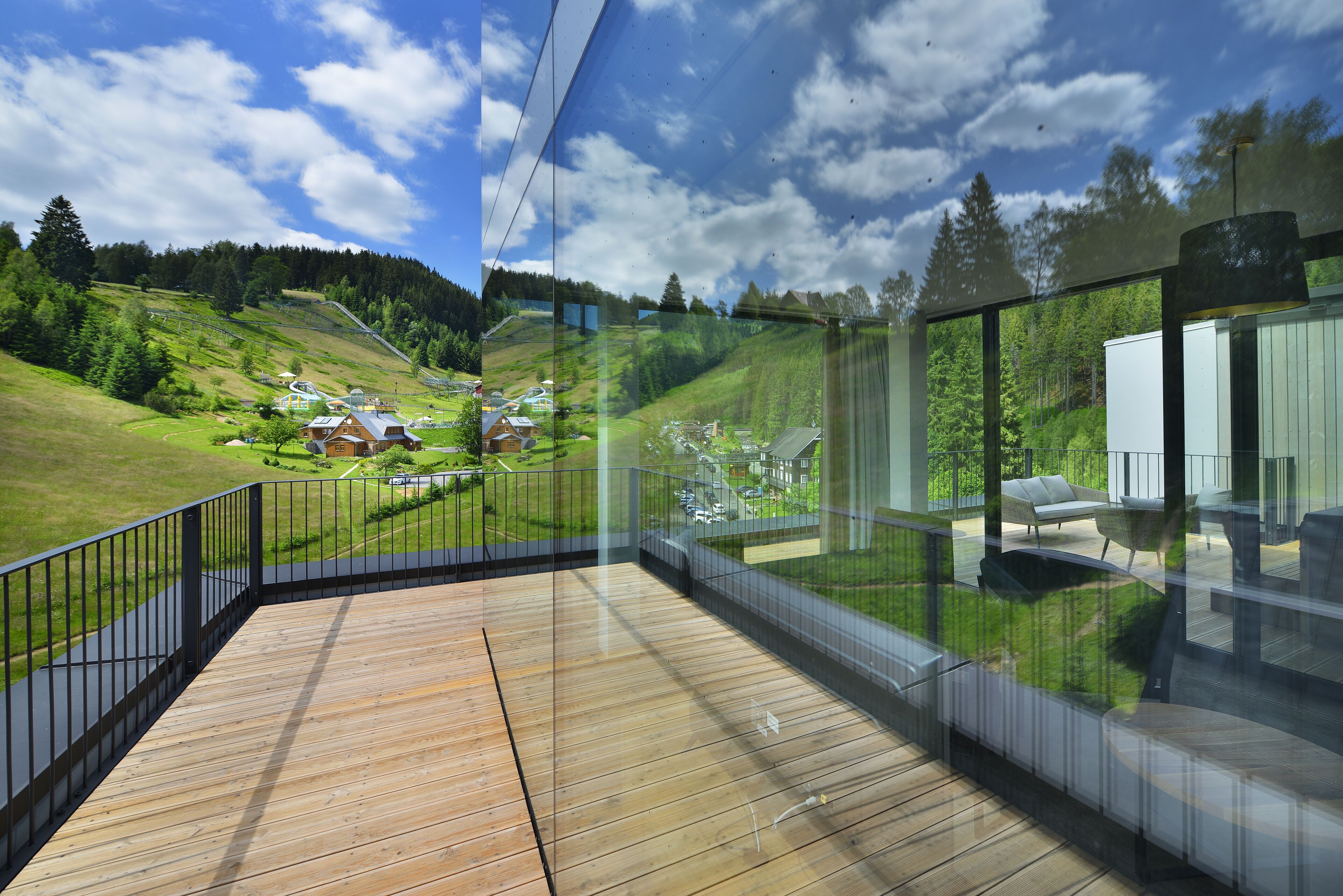 Ein moderner Glasbalkon mit Holzterrasse, der auf eine grüne Landschaft und einen klaren Himmel blickt.