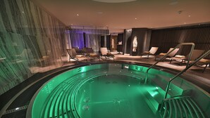 A modern indoor pool with a green-lit water feature, lounge chairs, and a spa area with a stone wall and a blue-lit partition. - Grand Hotel Hradec (Pec pod Snezkou)