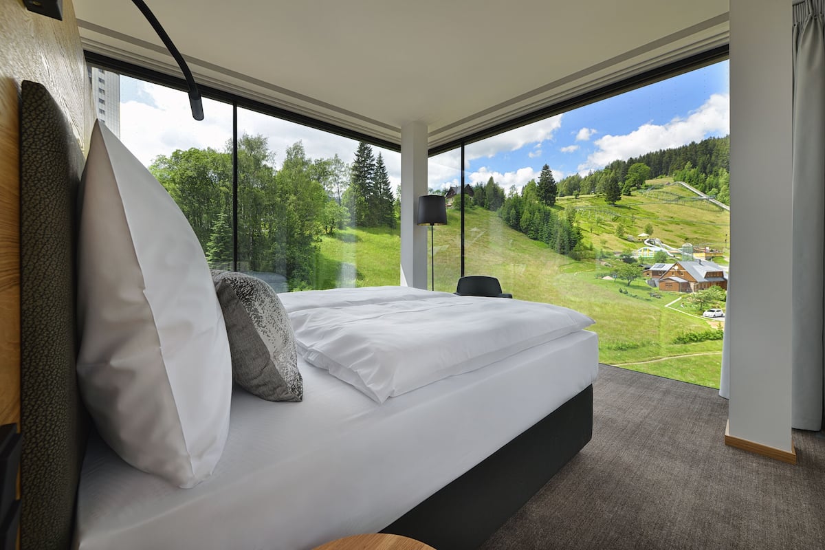 Ein modernes Hotelzimmer mit einem großen Bett, Blick auf eine grüne Landschaft und einem Fenster mit schwarzem Rahmen.