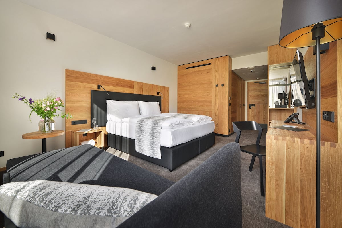 Ein modernes Hotelzimmer mit einem großen Bett, einem Schreibtisch, einem Sessel und einem kleinen Tisch mit Blumen in einer Vase.