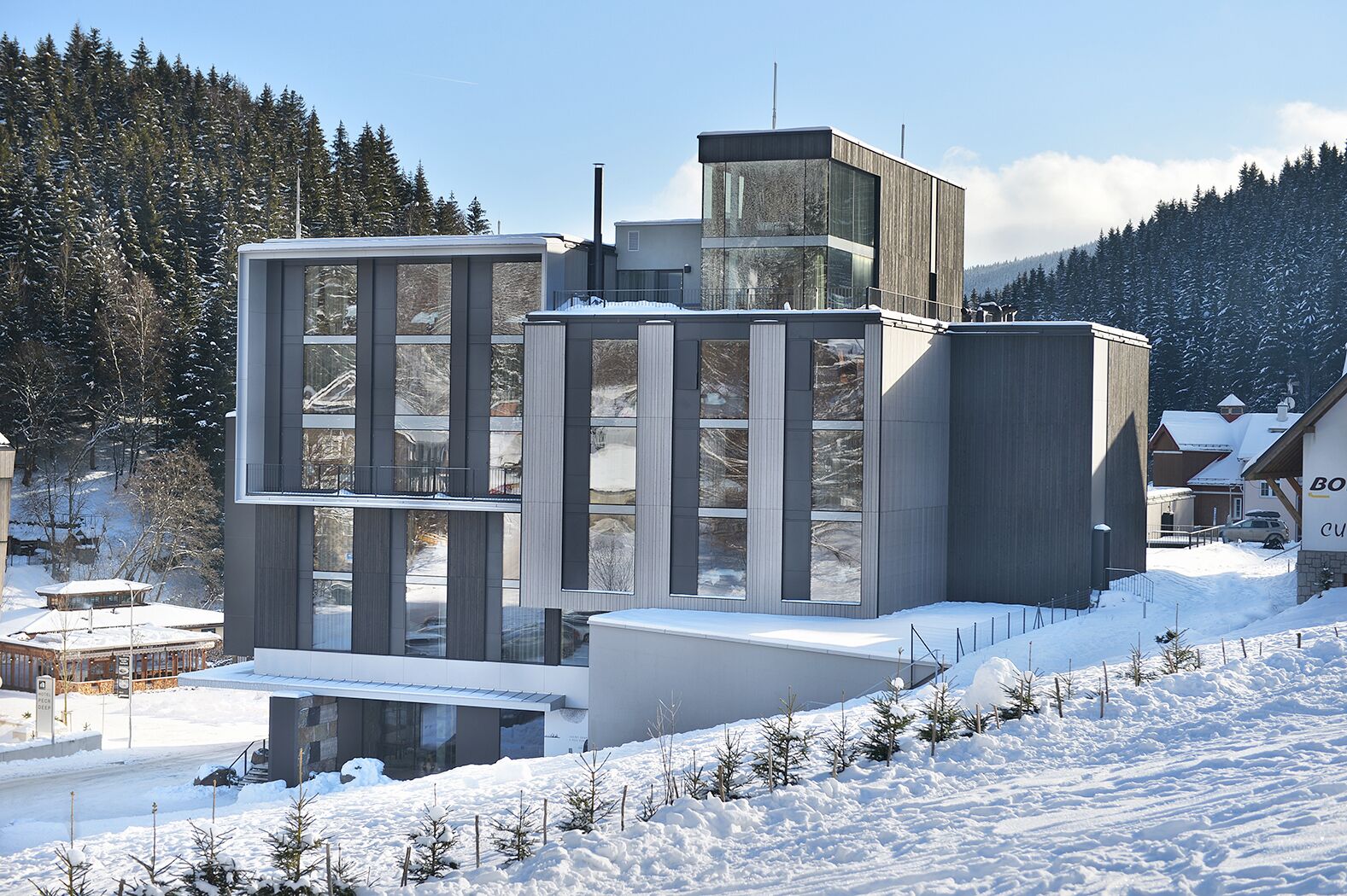 A modern, multi-story building with large windows and a flat roof, surrounded by snow-covered trees and a clear sky.