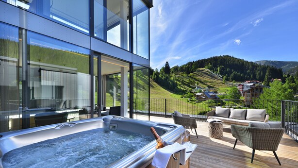 A hot tub on a wooden deck with a bottle of champagne and a glass, overlooking a scenic hillside with houses. - Grand Hotel Hradec (Pec pod Snezkou)