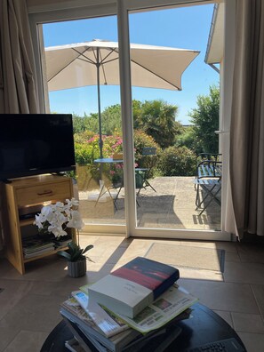 Una habitación con una televisión sobre un mueble de madera, una mesa con libros y una orquídea en maceta, y un ventanal corredizo que da a un patio exterior con mesa y sombrilla.