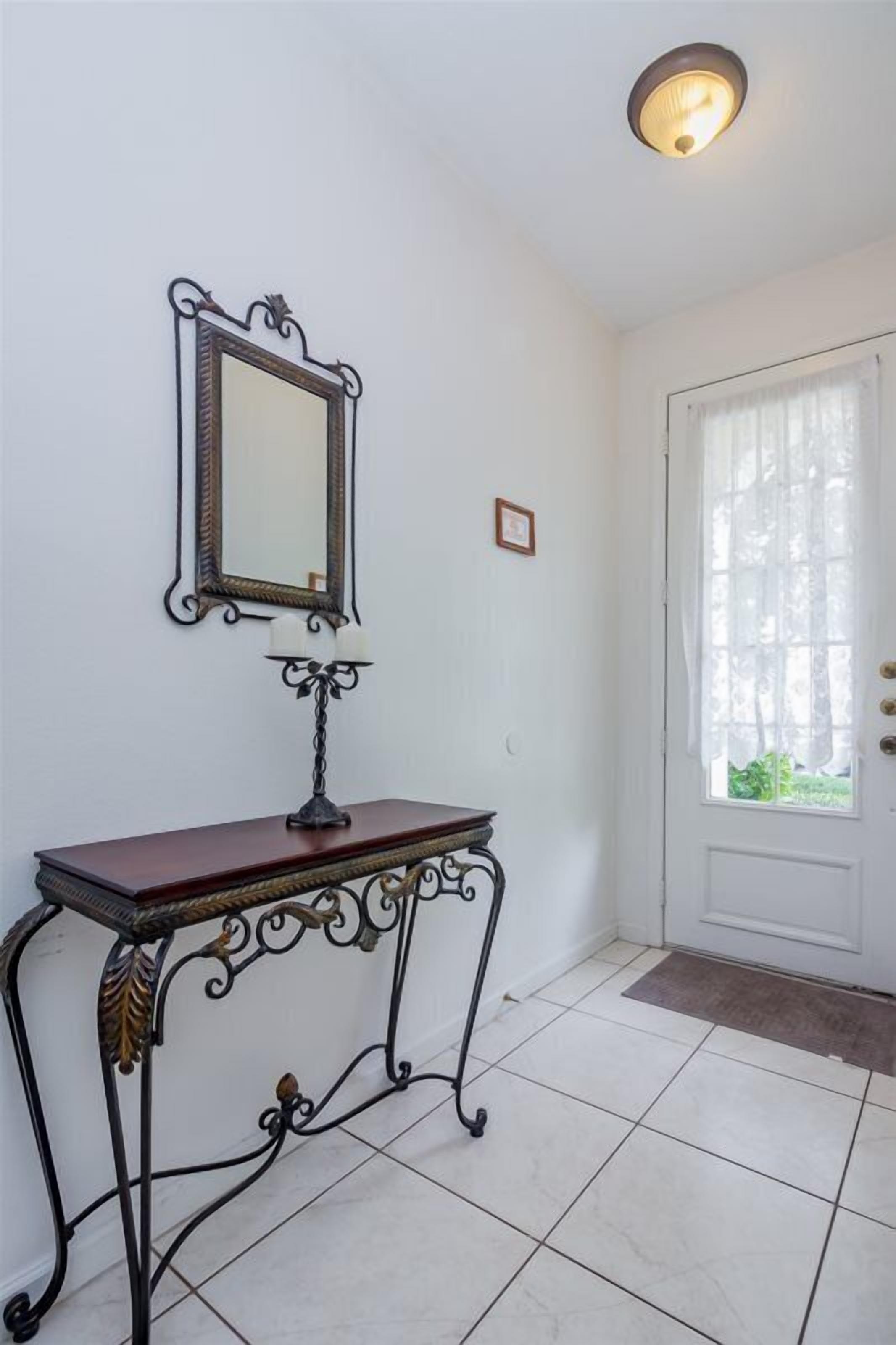 A room with a white door, a mirror on the wall, a small table with a lamp, and a window with curtains.