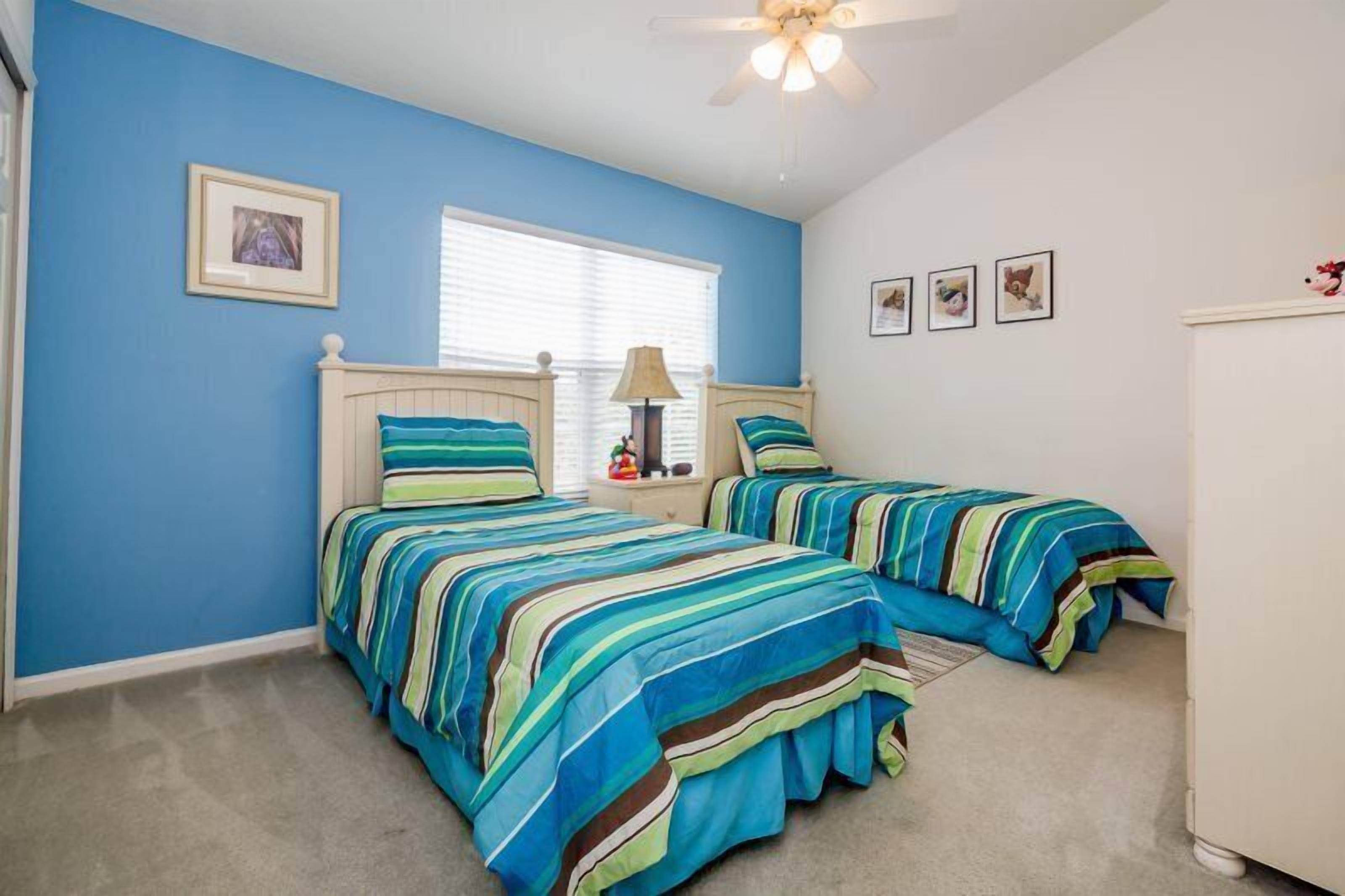 A bedroom with two beds, a ceiling fan, a window with blinds, and framed pictures on the wall.