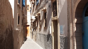 Une rue étroite, bordée de bâtiments de chaque côté, avec des portes cintrées et des fenêtres dotées de volets bleus.