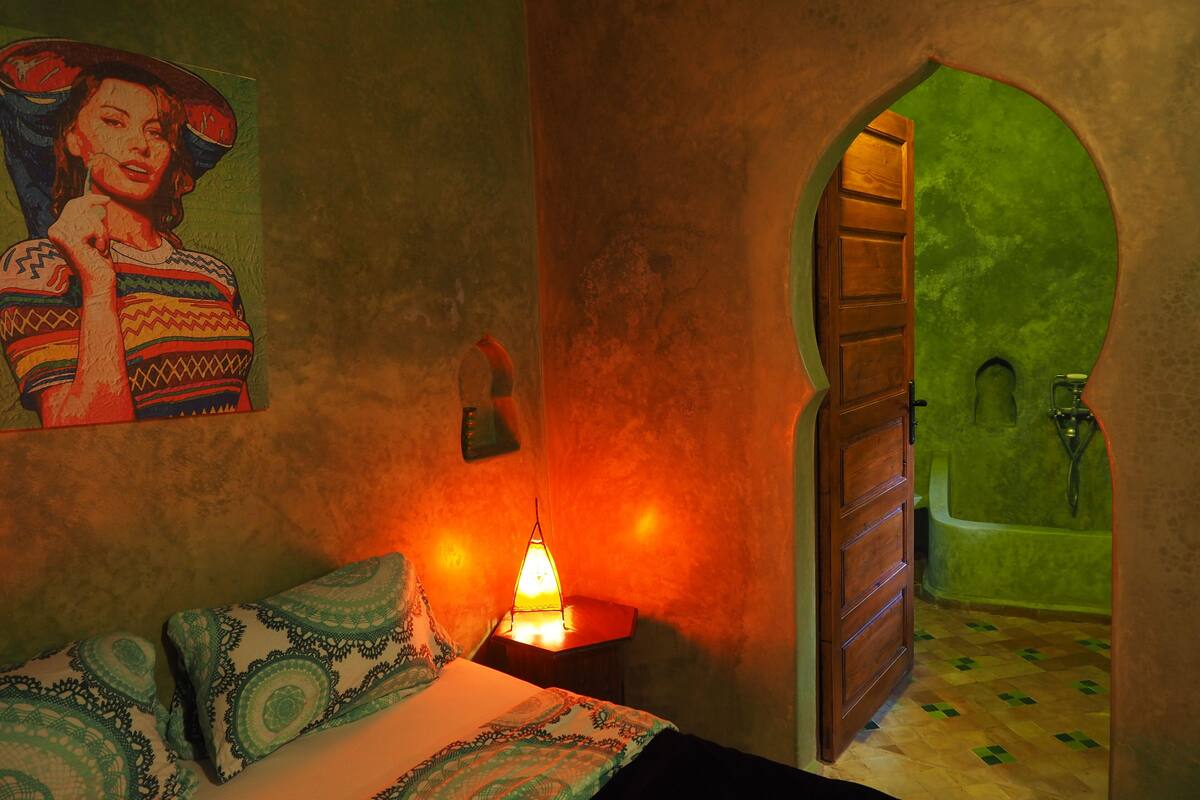 Une pièce comprenant un lit, une table de chevet avec une lampe, une arche verte menant à une salle de bain avec une baignoire, et un luminaire fixé au mur.