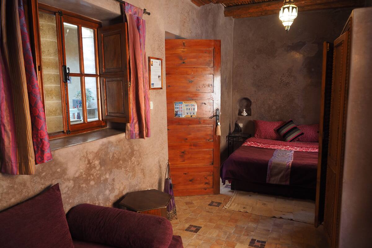 Une chambre confortable avec une porte en bois, un lit recouvert d’une couverture rouge, une petite table et une fenêtre avec des rideaux.