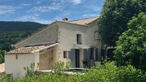 A rustic building with a tiled roof and a small porch, surrounded by lush greenery and a clear blue sky. - Provencal farmhouse with pool at the foot of Mont-Ventoux (Malaucène)
