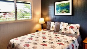 Um quarto com cama, ventilador de teto, janela com persianas, mesa de cabeceira, abajur e um quadro na parede.