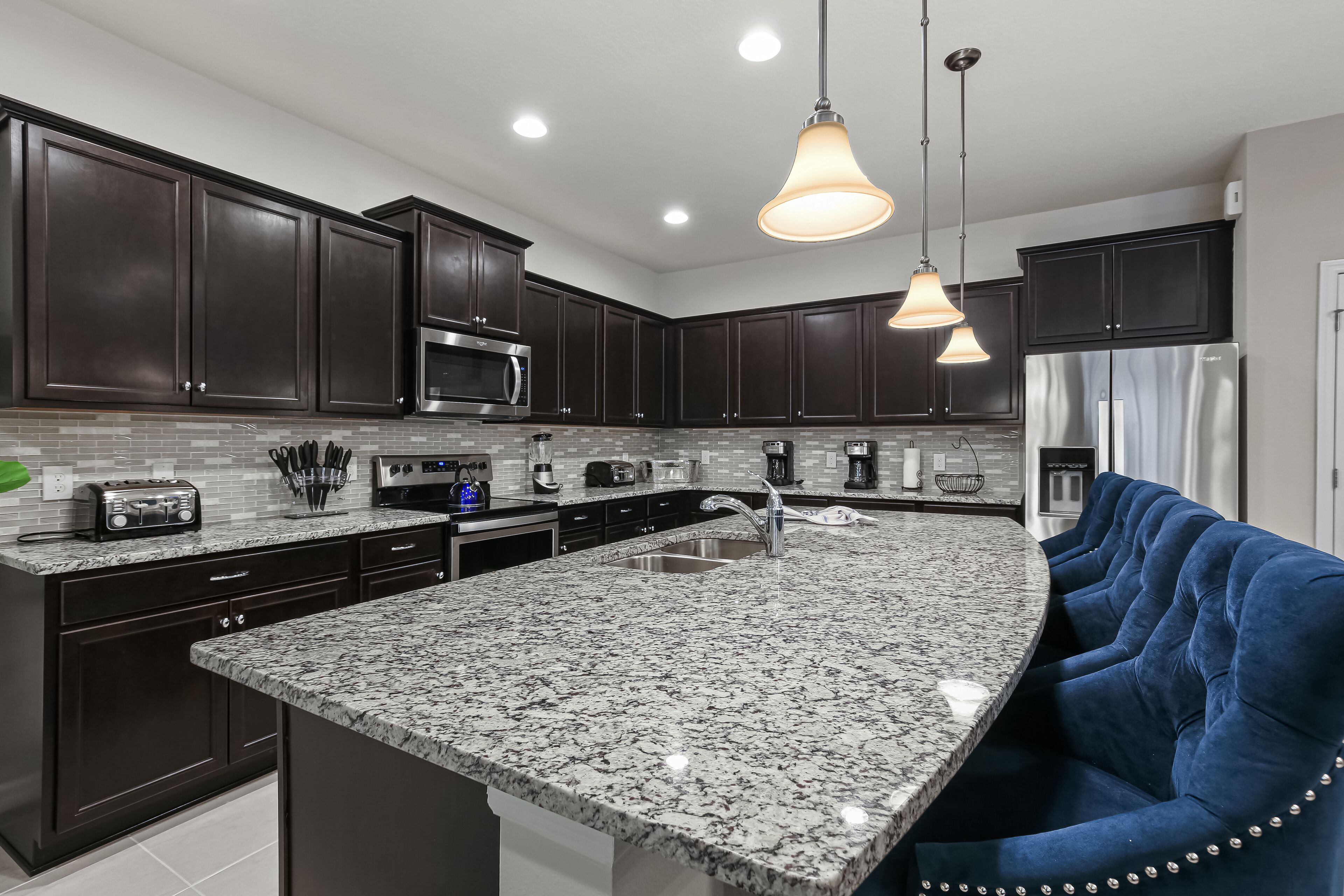 Cocina moderna con armarios oscuros, electrodomésticos de acero inoxidable, encimera de granito y sillas tapizadas en azul.