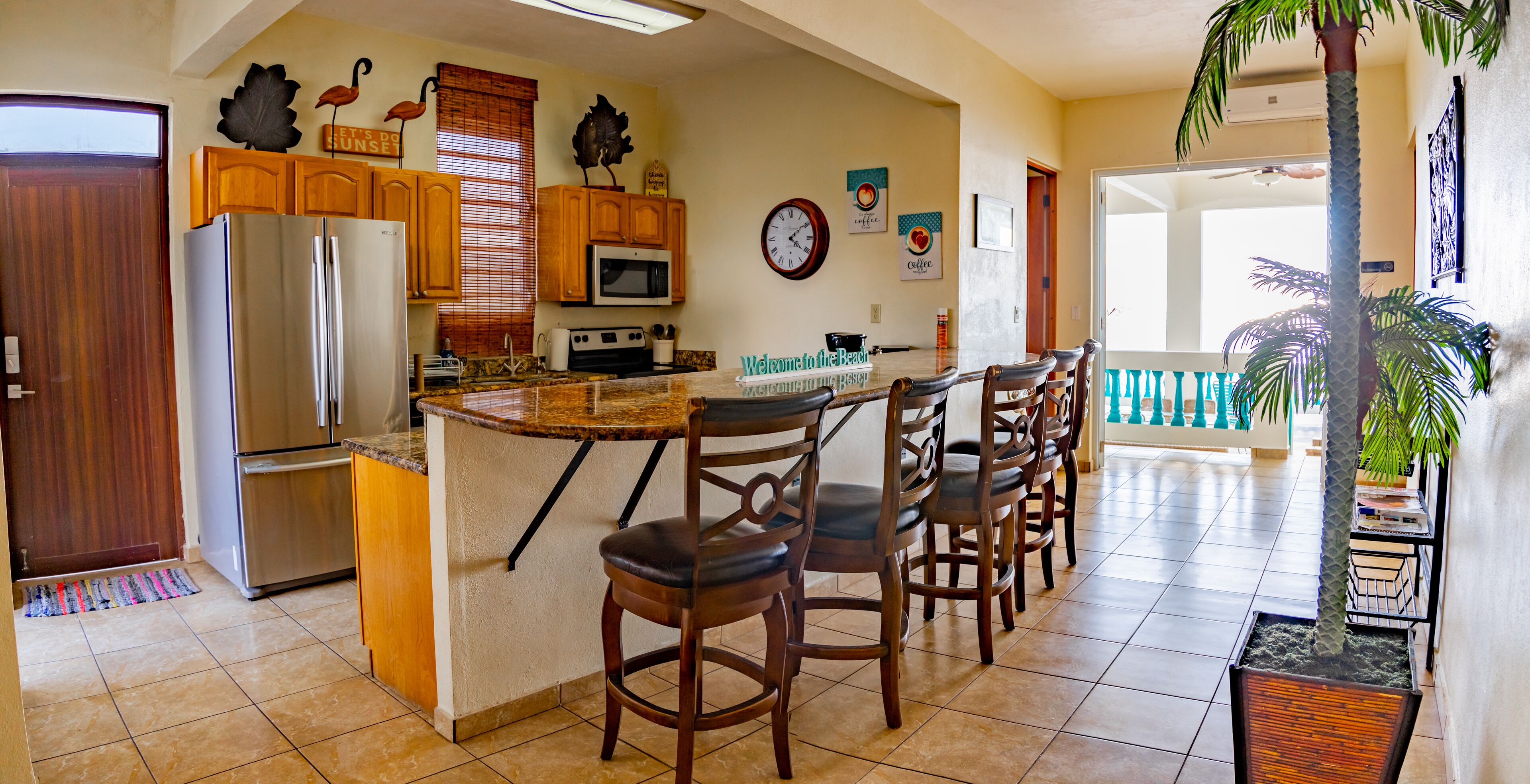A kitchen with a central island, wooden cabinets, a refrigerator, and a microwave. There are chairs around the island and a clock on the wall.