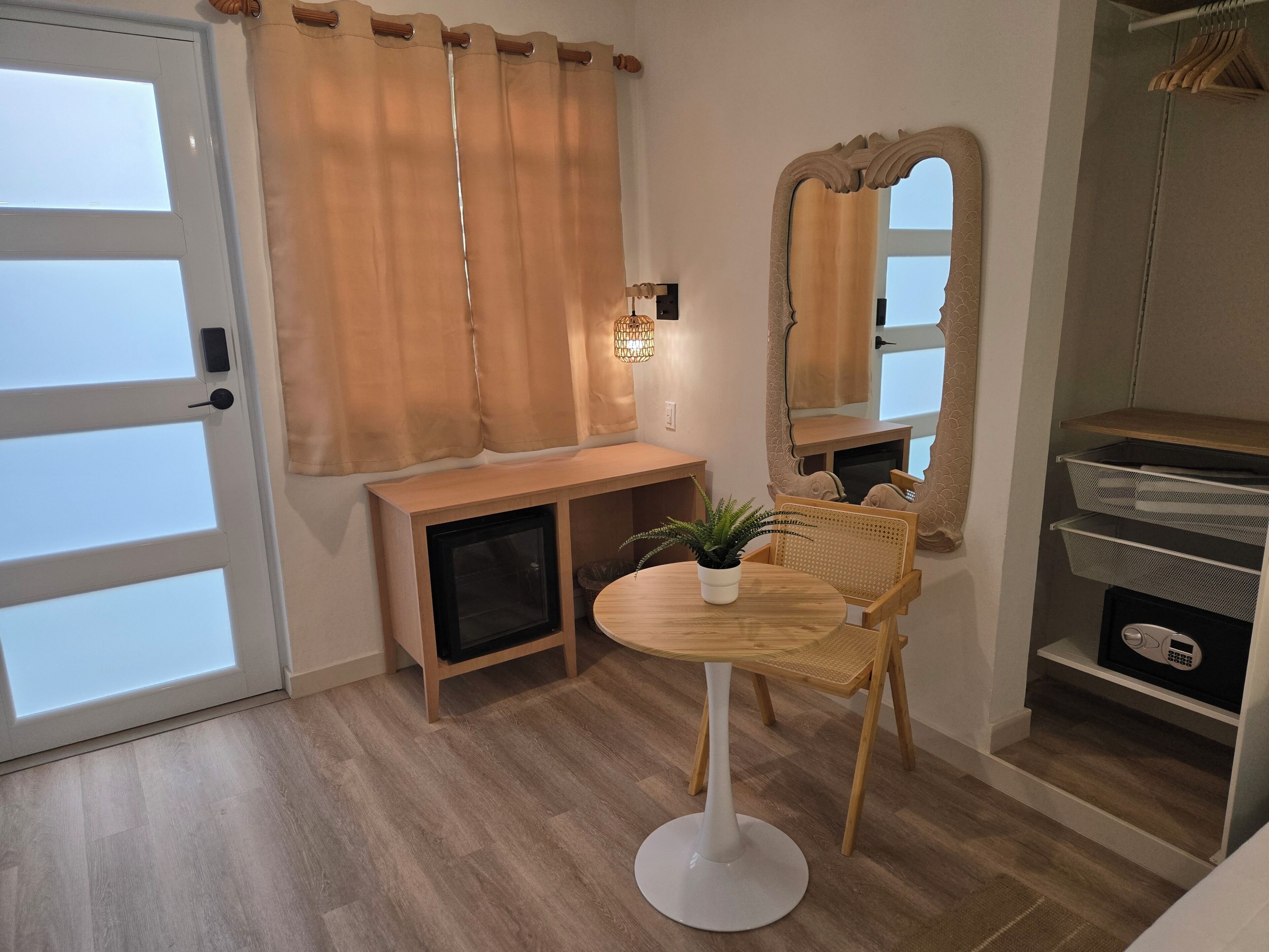 A room with a wooden floor, a white pedestal table with a potted plant, a wicker chair, a wooden console table, a mirror with a decorative frame, and a clothing rack.