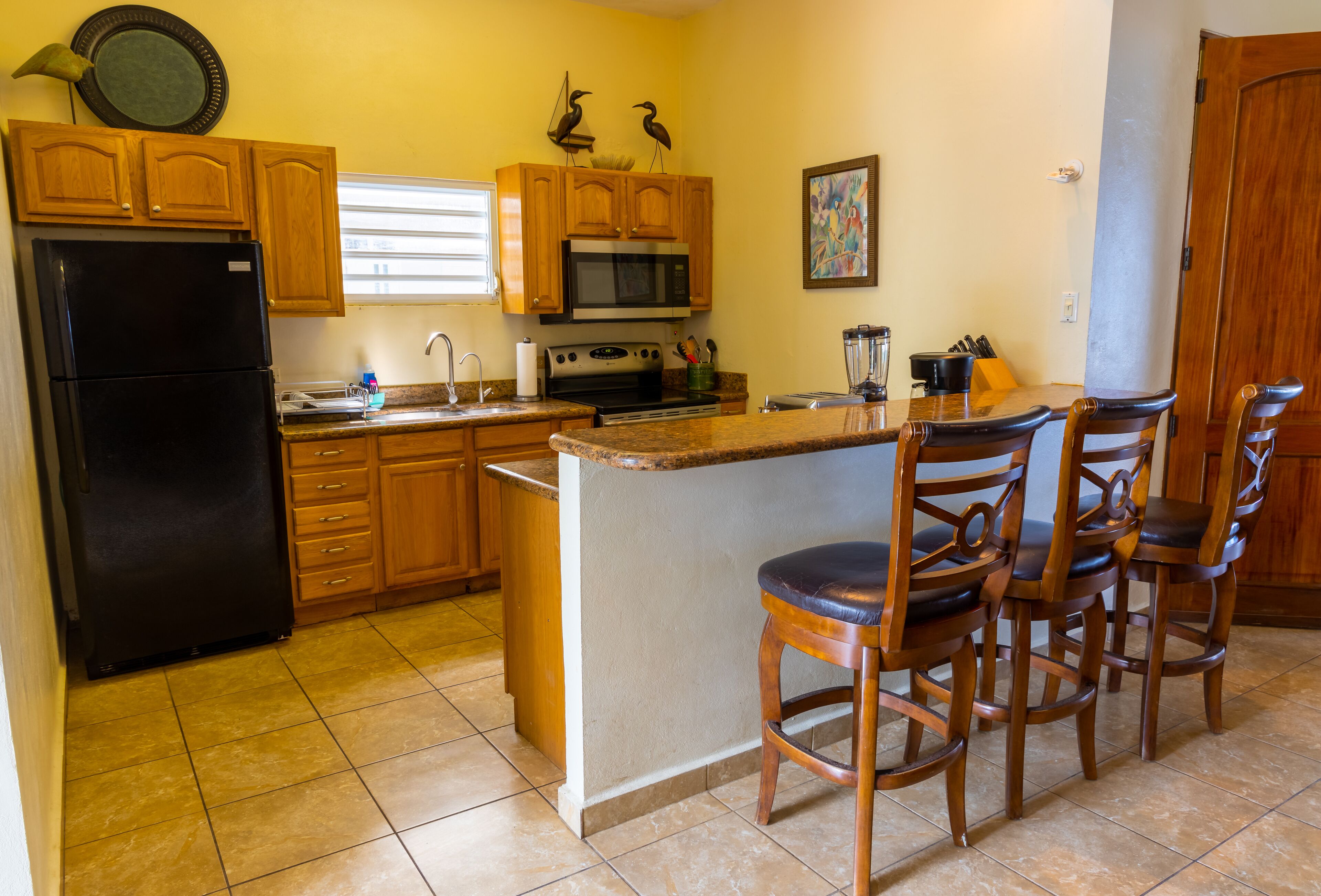 Cocina con armarios de madera, un refrigerador negro, microondas, horno, lavaplatos y una barra con sillas de madera.