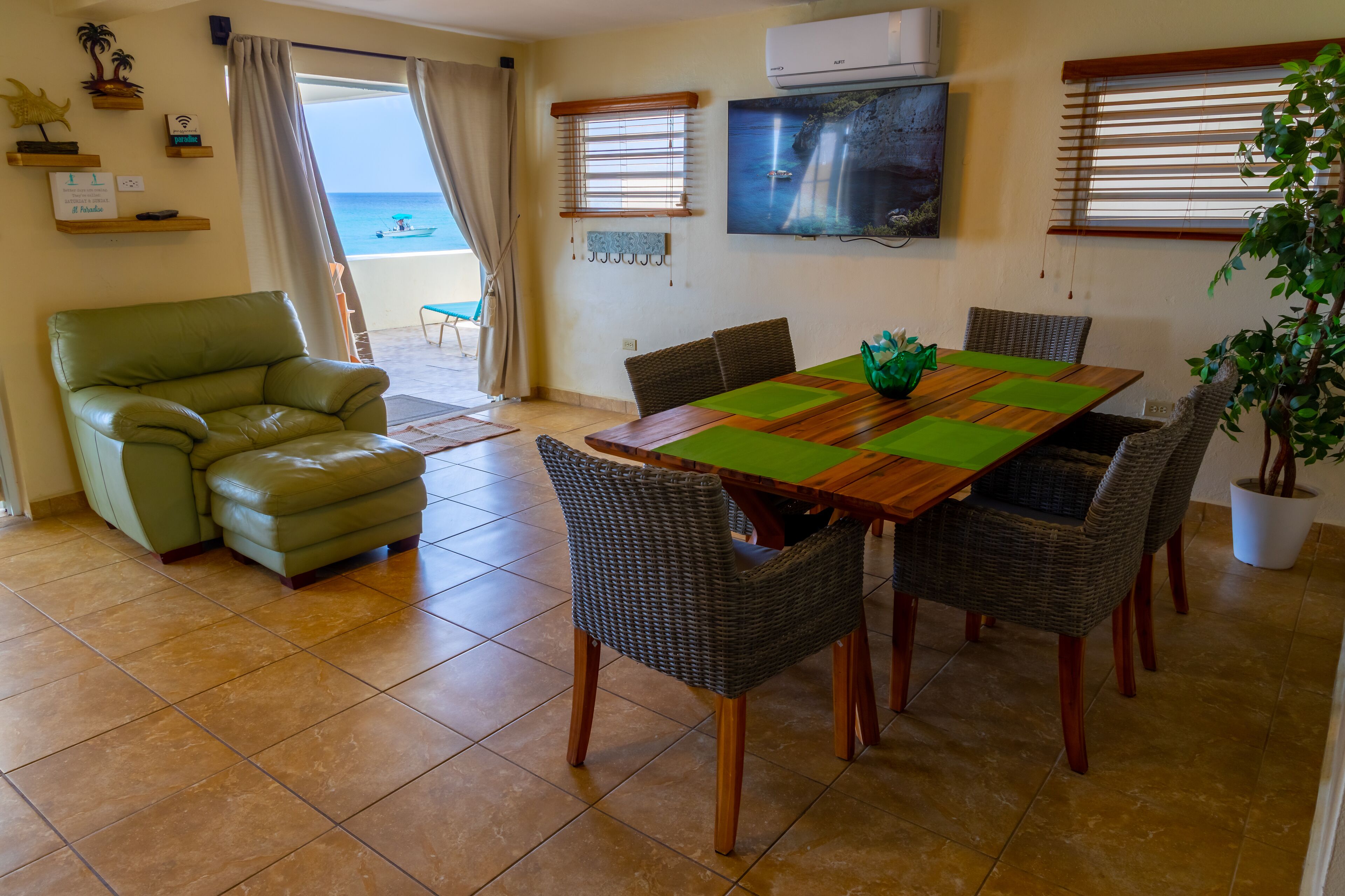 A living room with a green sofa, a wooden dining table with green placemats, wicker chairs, a potted plant, and a view of the sea through a window.