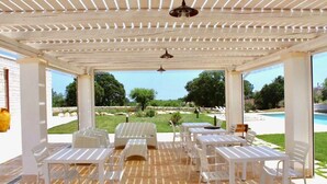 Un espacio al aire libre cubierto con mesas y sillas blancas, suelo de madera y vista a una piscina y jardín.
