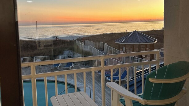 Un balcón con vista a la playa al atardecer, una piscina y un pabellón.