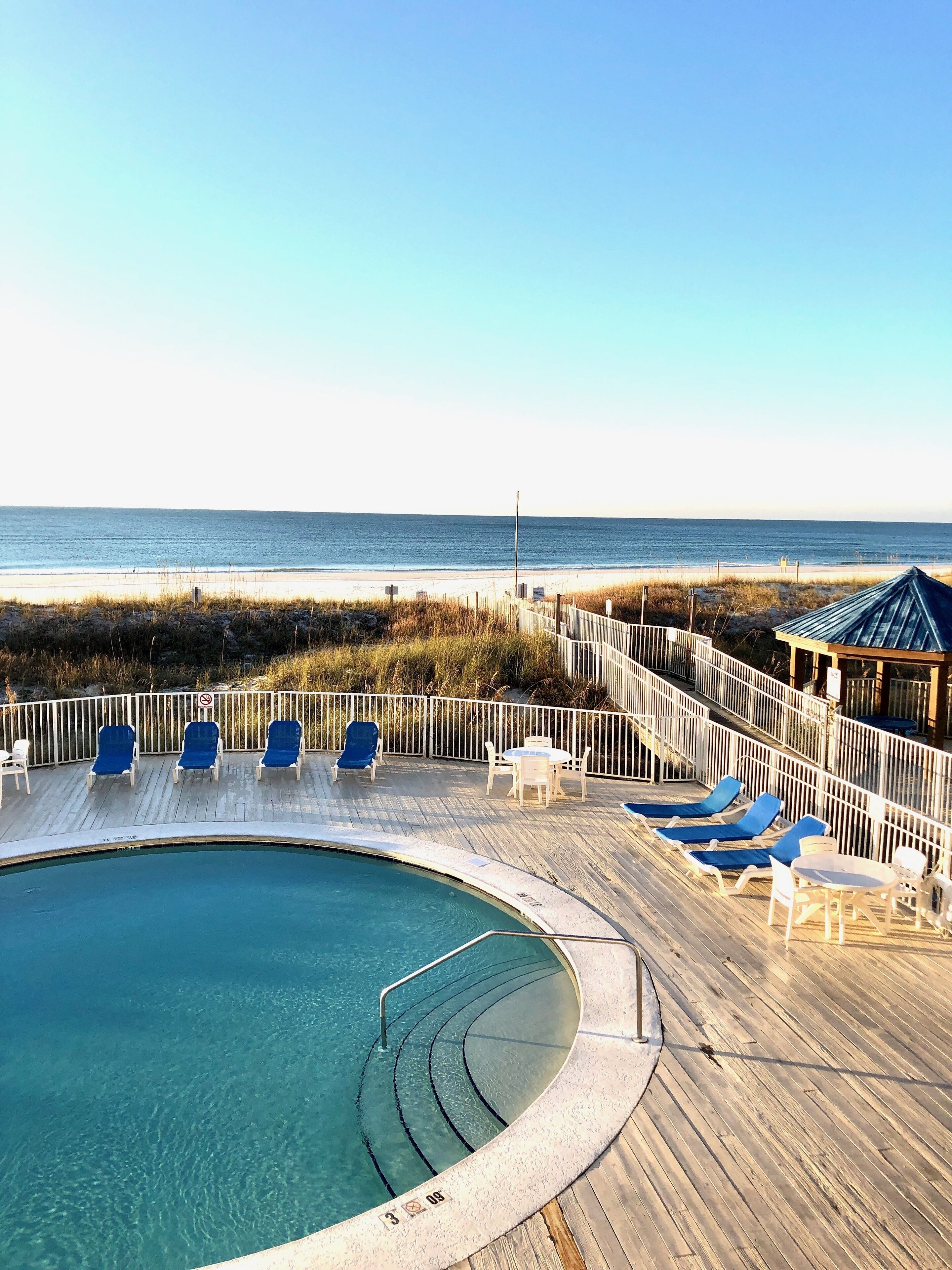 A pool area with a wooden deck, blue lounge chairs, and a view of the beach.