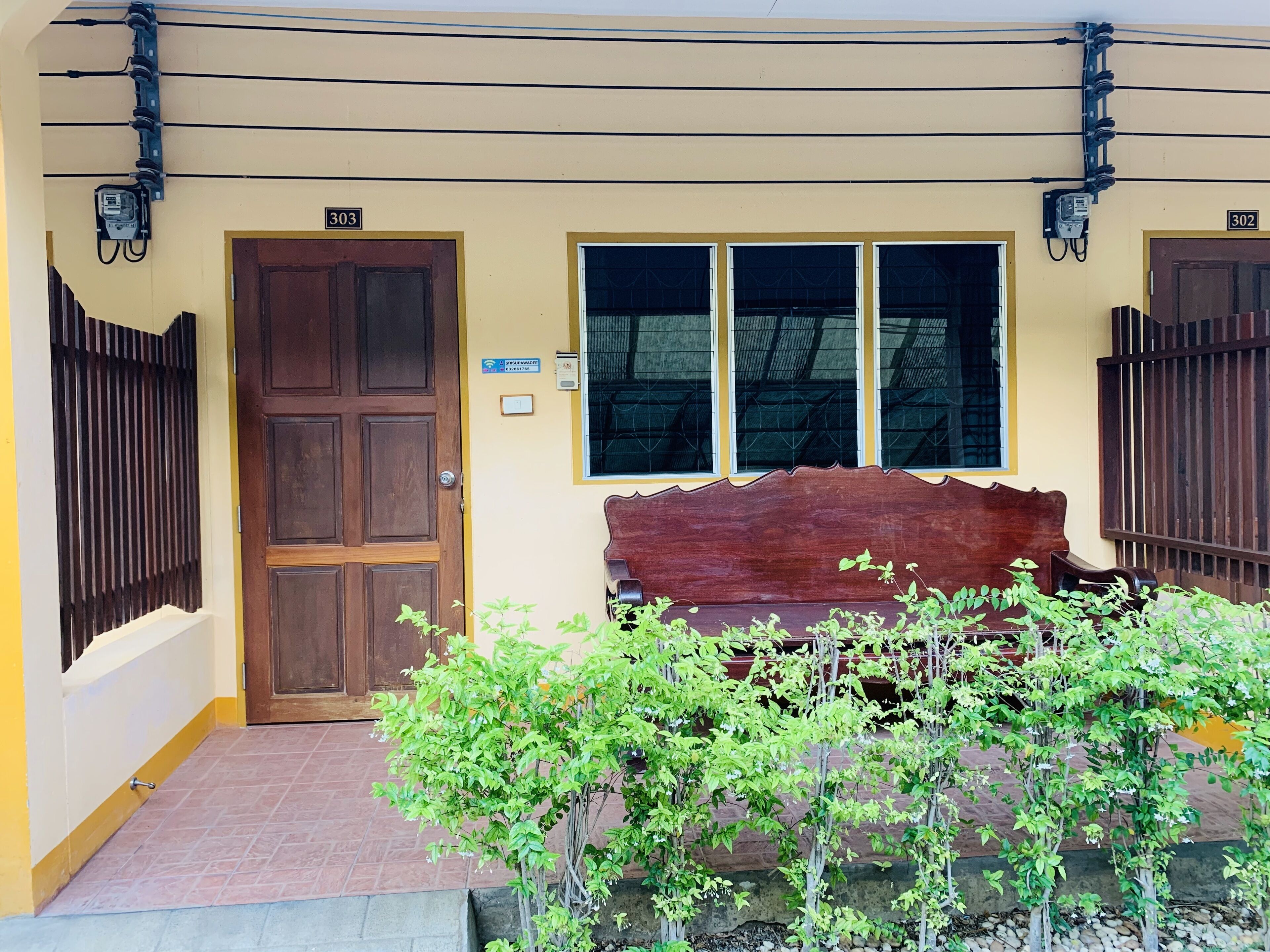 A building entrance with a wooden door labeled '303', a bench, and a small garden area with green plants.